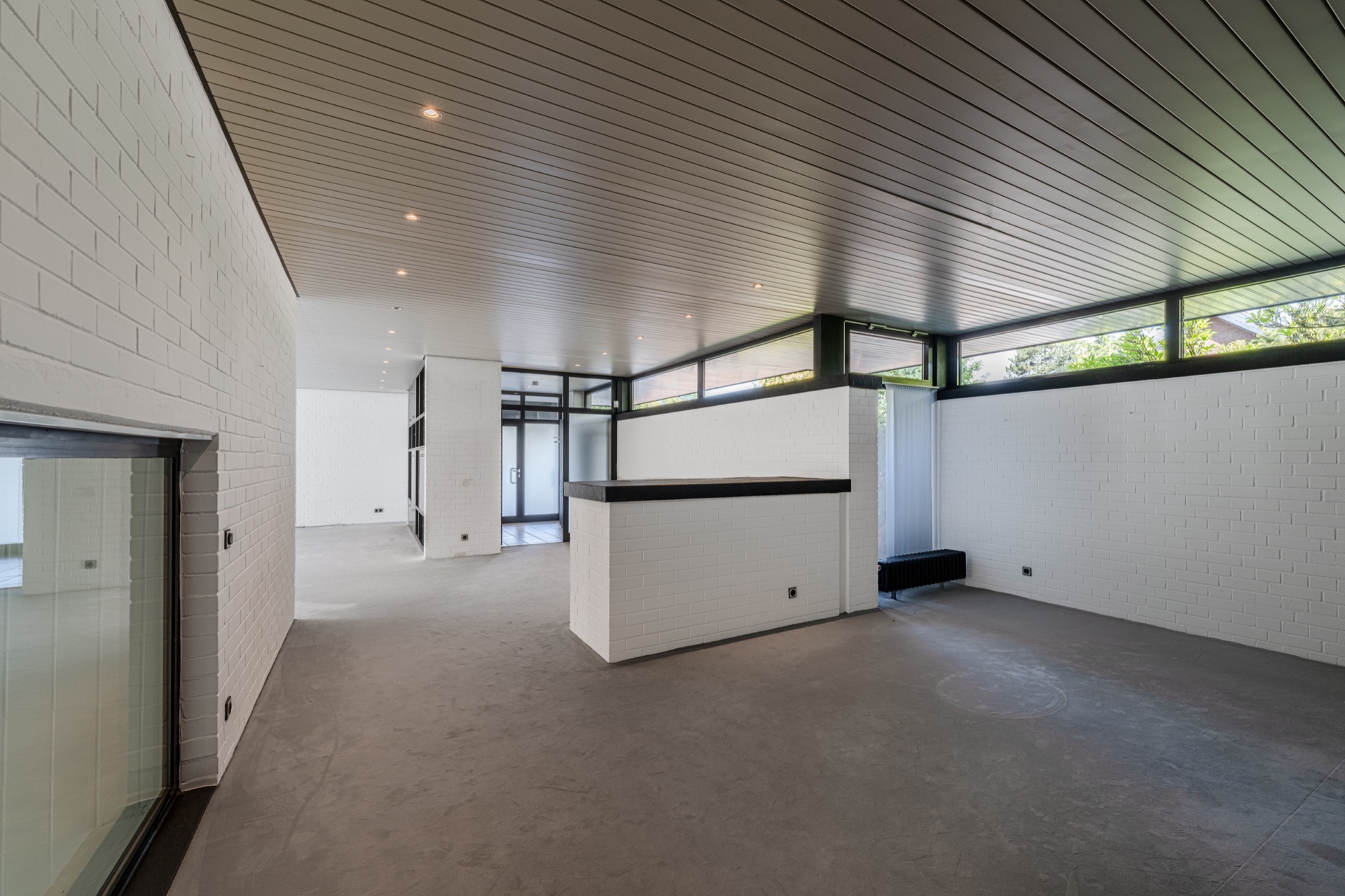 Großer, leerer Raum mit weißen Wänden, Fenstern und grauem Boden in einem Wohnhaus.