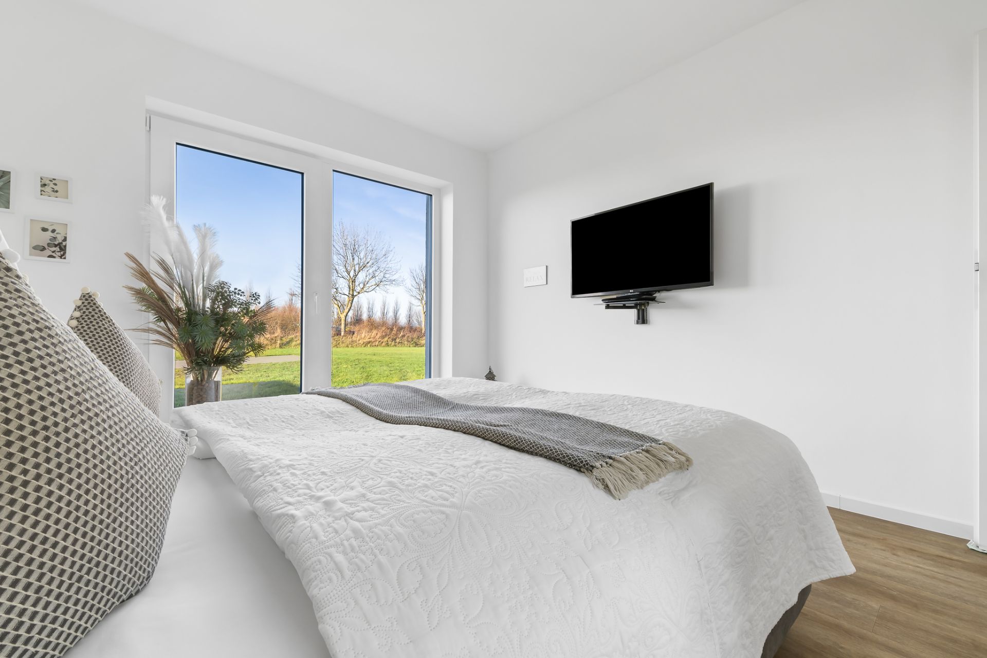 Helles Schlafzimmer mit Doppelbett, Fernseher an der Wand und Blick auf den Garten durch große Fenster.