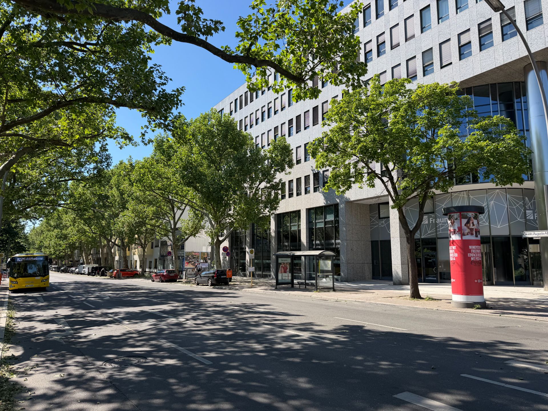 Großes Bürogebäude mit vielen Fenstern, Bäumen, Bushaltestelle und Straße im Vordergrund.