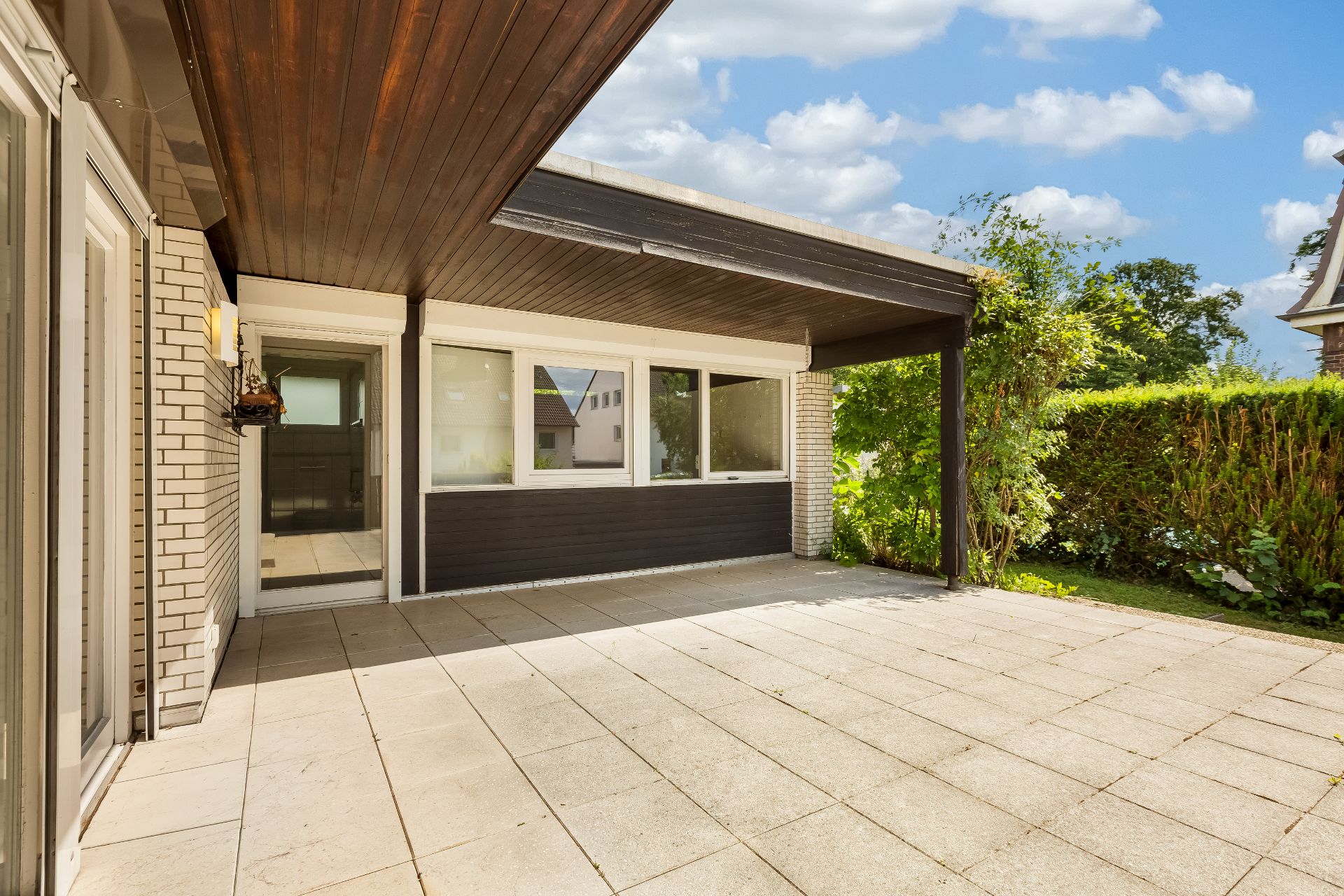 Überdachte Terrasse mit Fliesenboden, weißen Wänden und großen Fenstern.