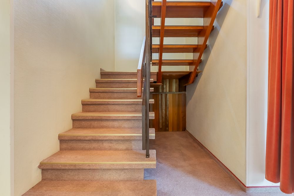 Holztreppe in einem Flur mit beige Wand und rotem Vorhang rechts.