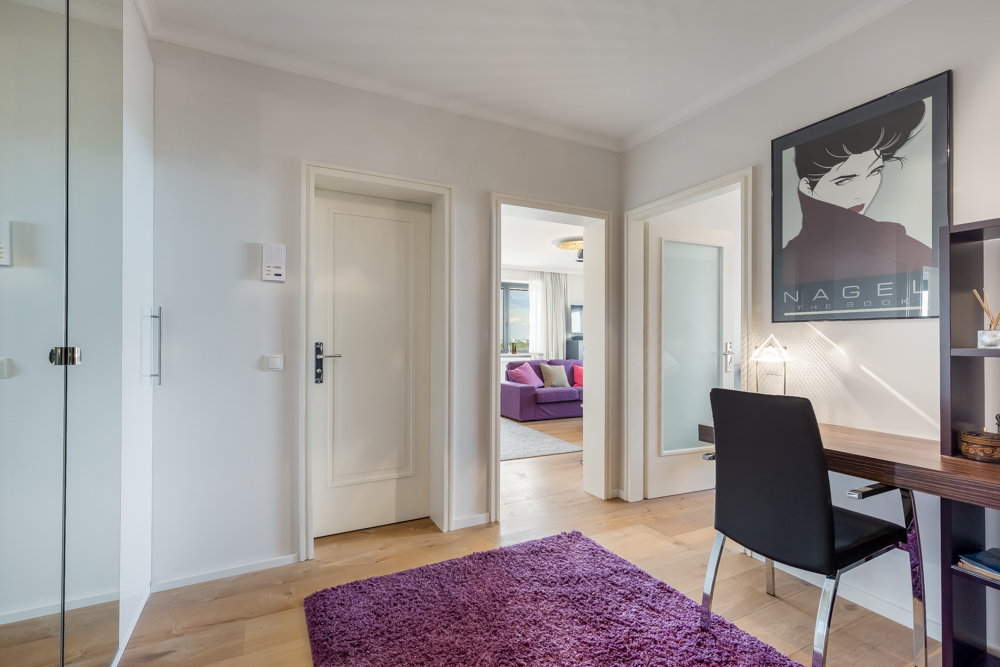 Heller Flur mit Holzboden, Schreibtisch, lila Teppich und Blick ins Wohnzimmer mit Sofa.
