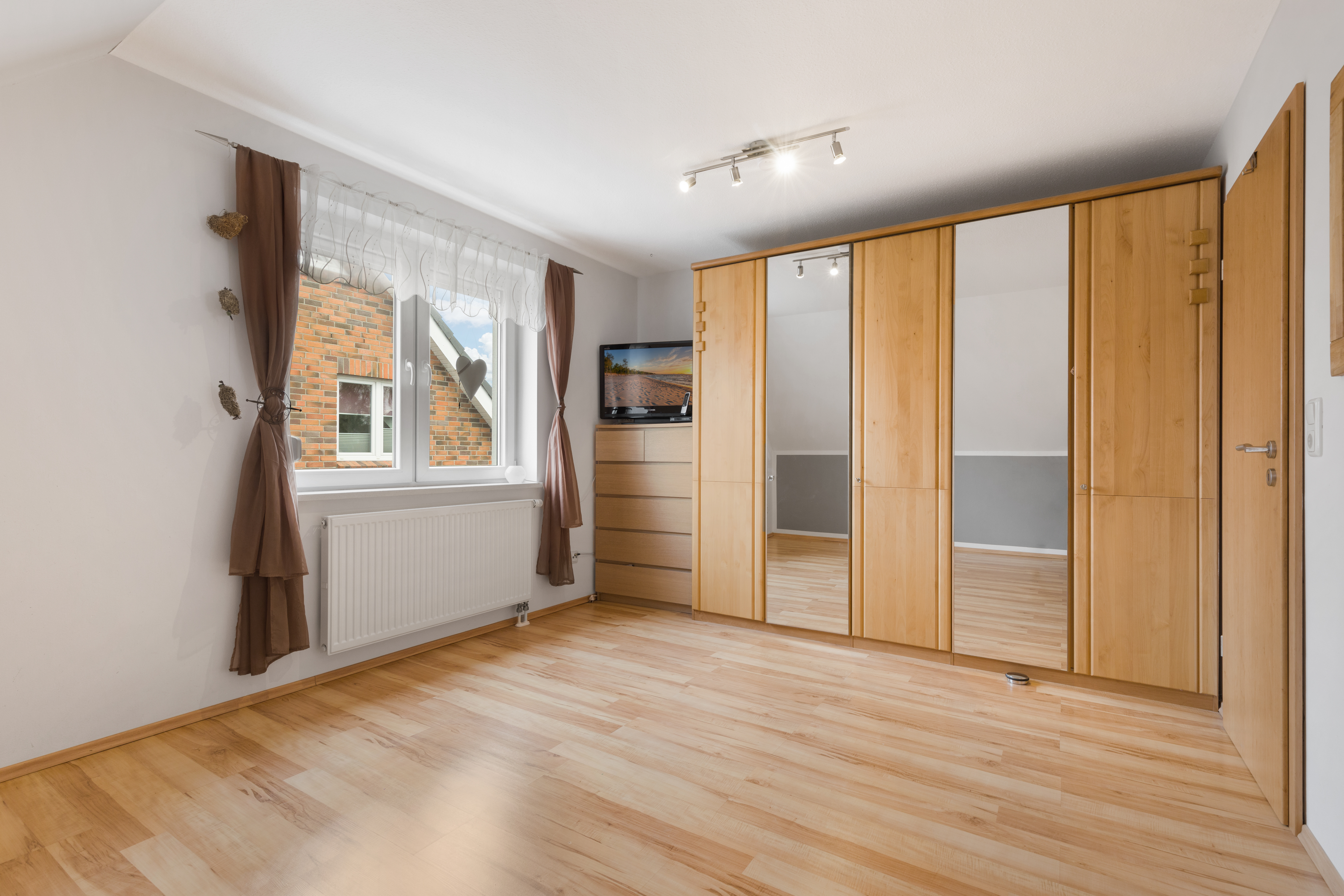 Helles Zimmer mit Holzfußboden, Kleiderschrank, Fenster mit Vorhängen und Kommode mit Fernseher.