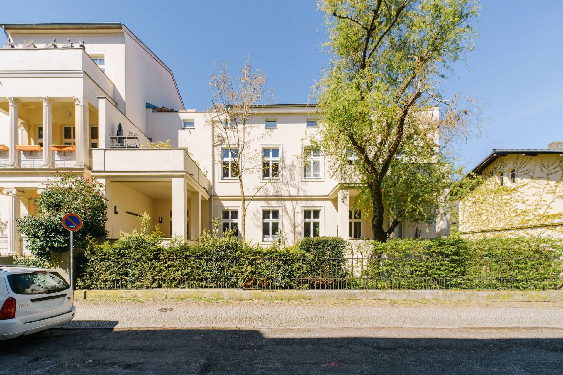 Helles Wohnhaus mit mehreren Fenstern, Balkon und Bäumen davor, im Sonnenschein.