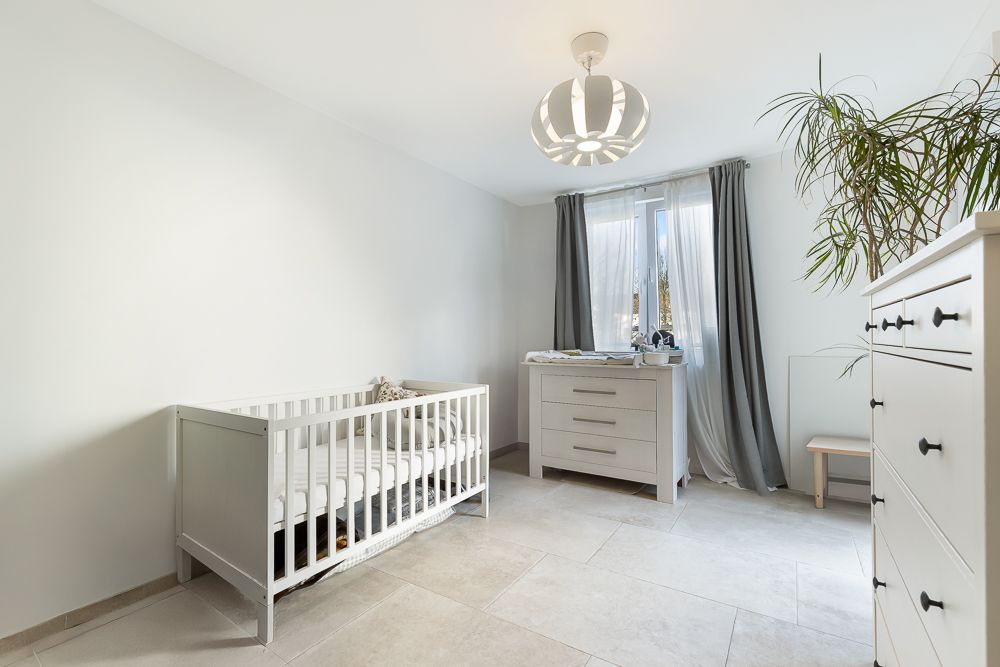 Helles Kinderzimmer mit Gitterbett, Kommode, Schrank und Pflanze am Fenster.