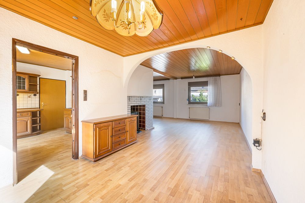 Helles Wohnzimmer mit Holzdecke, Parkettboden, Durchgang zur Küche und großen Fenstern.