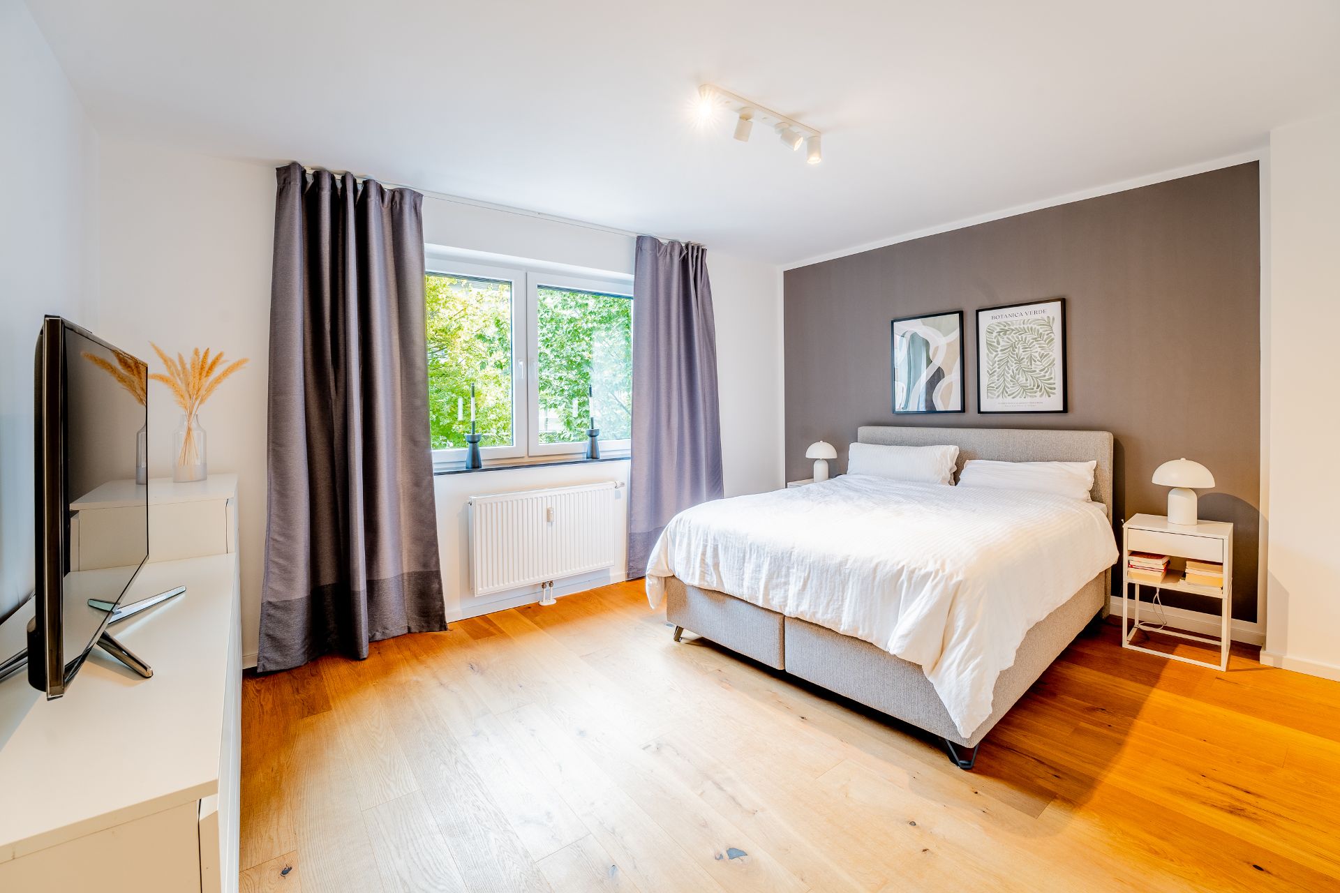 Helles Schlafzimmer mit Doppelbett, Fernseher, Holzboden und zwei Fenstern mit grauen Vorhängen.