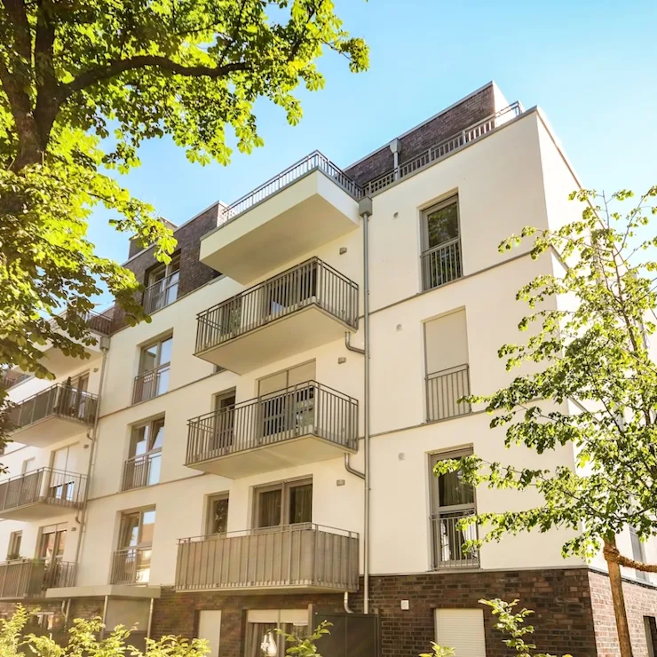 Modernes Wohnhaus mit mehreren Balkonen, umgeben von Bäumen, bei Sonnenschein.