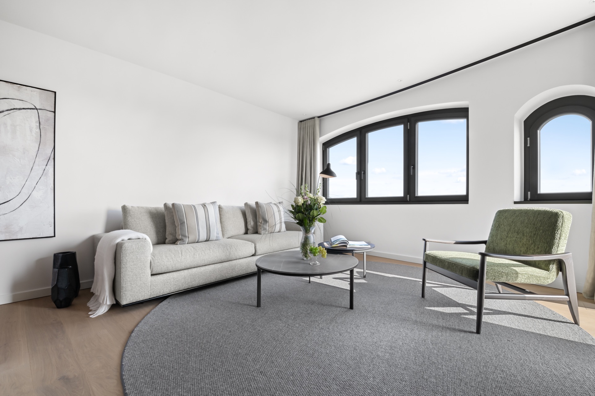 Modernes Wohnzimmer mit Sofa, grünem Sessel, rundem Teppich und großen Fenstern.