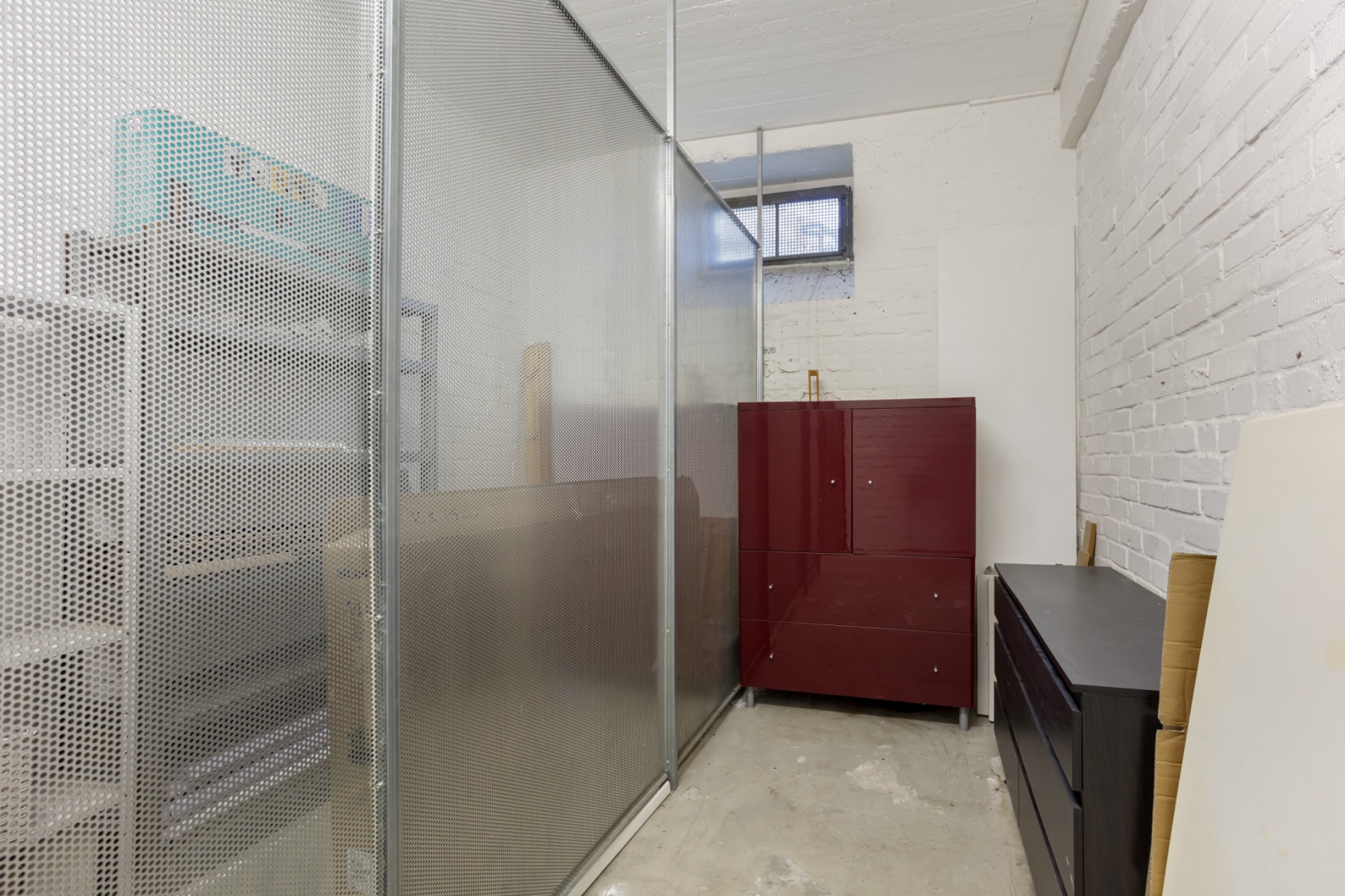Kellerraum mit Metallregal, rotem Schrank und schwarzer Kommode. Fenster an der Rückwand.