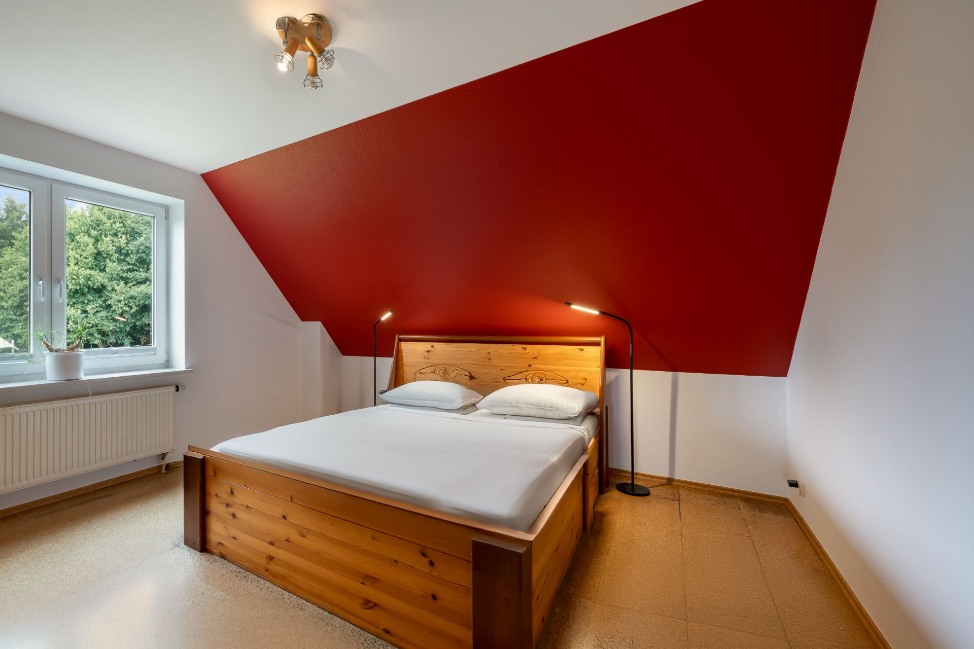 Schlafzimmer mit Holzbett, zwei Stehlampen und roter Dachschräge, Fenster auf der linken Seite.