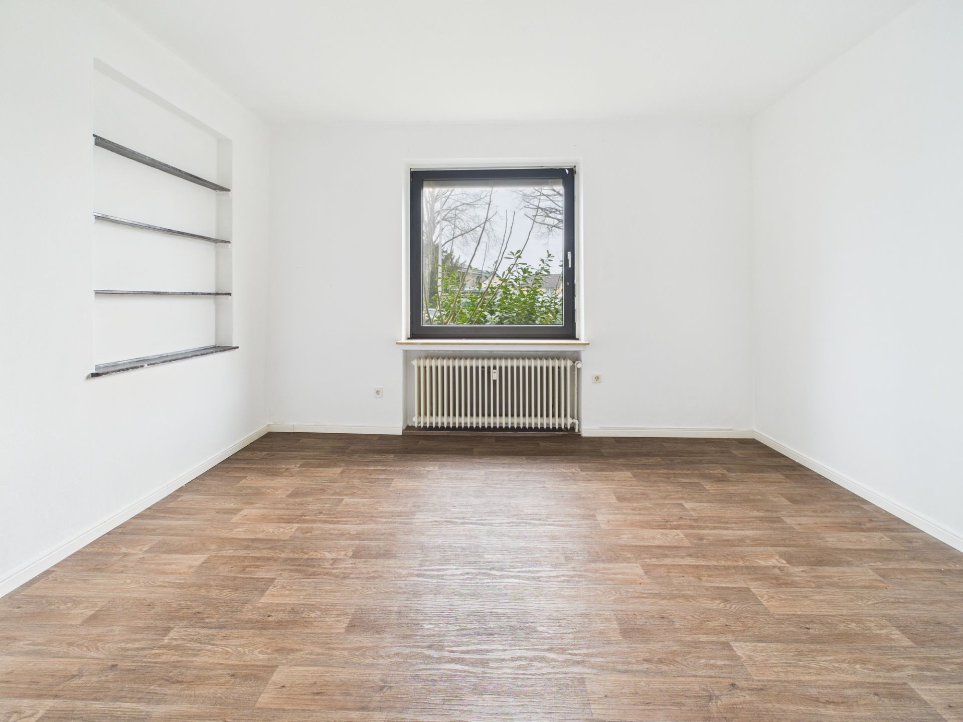 Leerer Raum mit Holzfußboden, einem Fenster und eingebauten Wandregalen links.