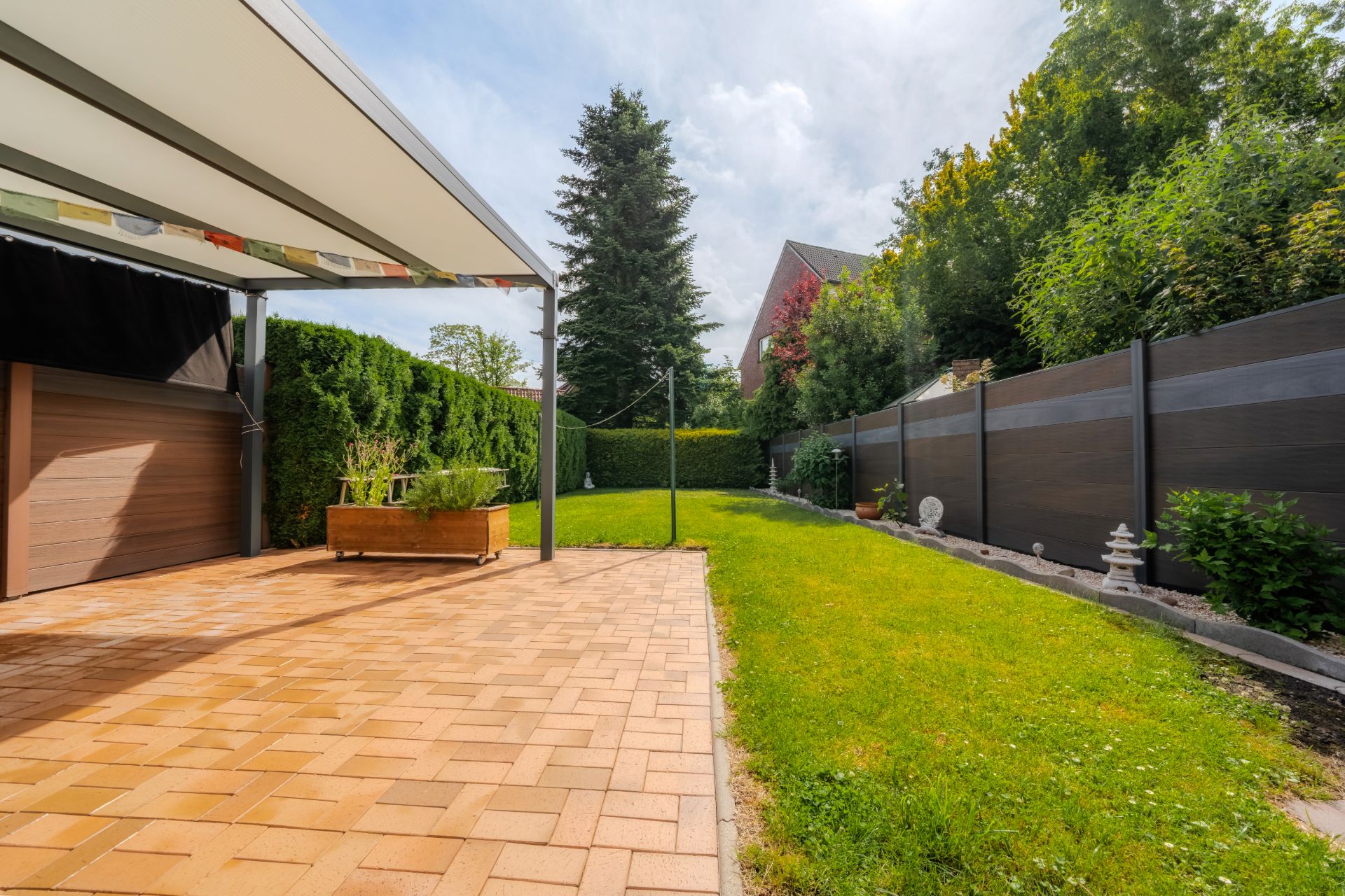 Gepflasterte Terrasse mit Überdachung und angrenzendem Garten, umgeben von Hecken und Zaun.