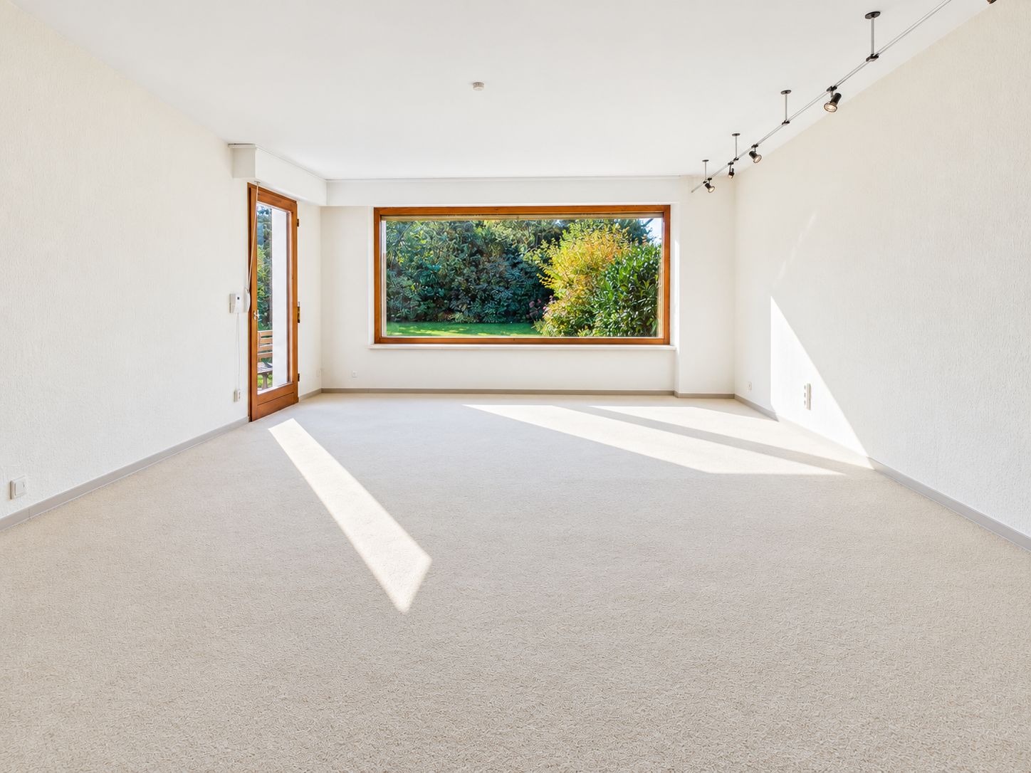 Leeres, helles Wohnzimmer mit großem Fenster und Tür zum Garten.