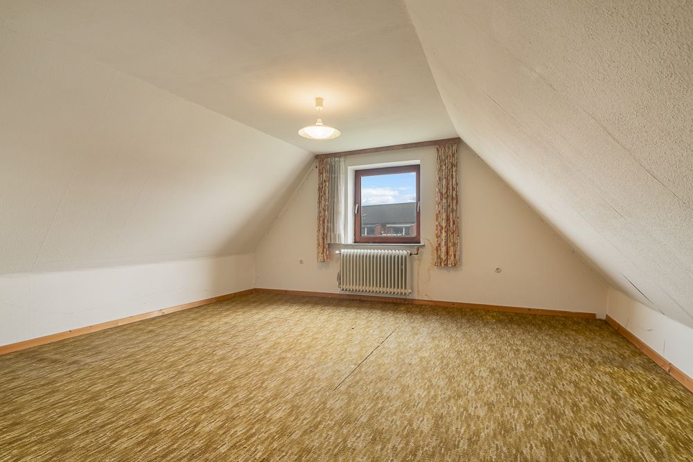 Leeres Dachzimmer mit Teppichboden, Fenster, Heizkörper und schrägen Wänden.