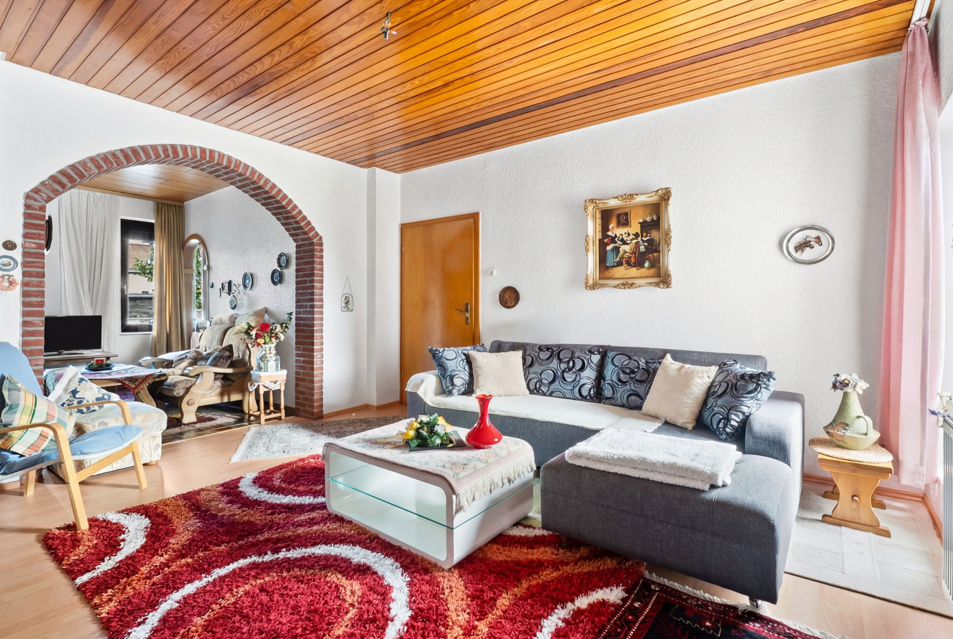Wohnzimmer mit grauem Sofa, rotem Teppich, Glastisch und Holzbalkendecke.