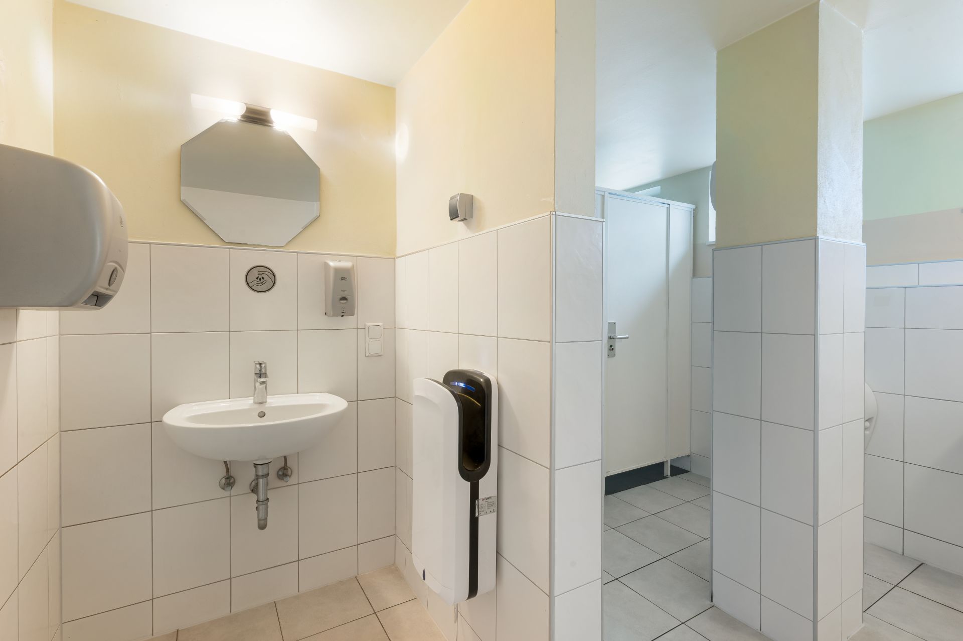 Helles Badezimmer mit Waschbecken, Seifenspender, Spiegel und einzelner Kabine im Hintergrund.