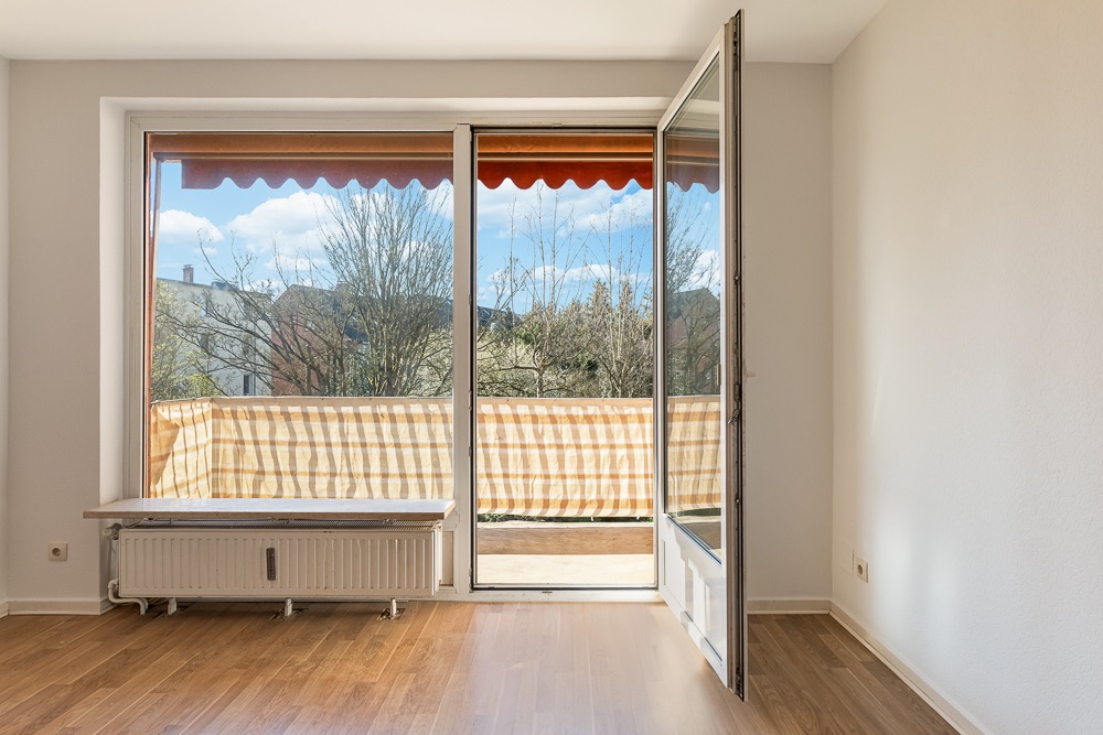 Helles Zimmer mit Holzfußboden und Zugang zu einem Balkon mit Markise und Ausblick ins Grüne.