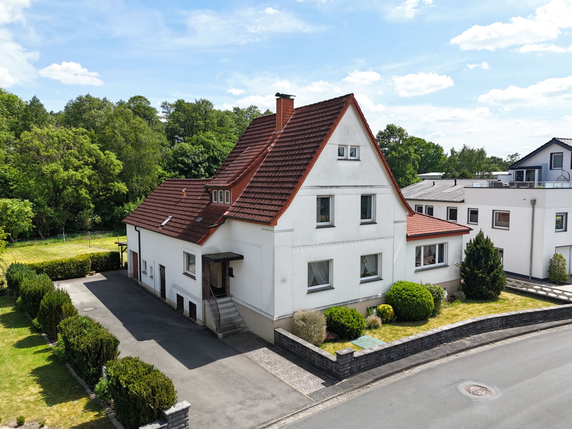Zweistöckiges Einfamilienhaus mit rotem Satteldach, Vorgarten und Einfahrt, umgeben von Büschen.