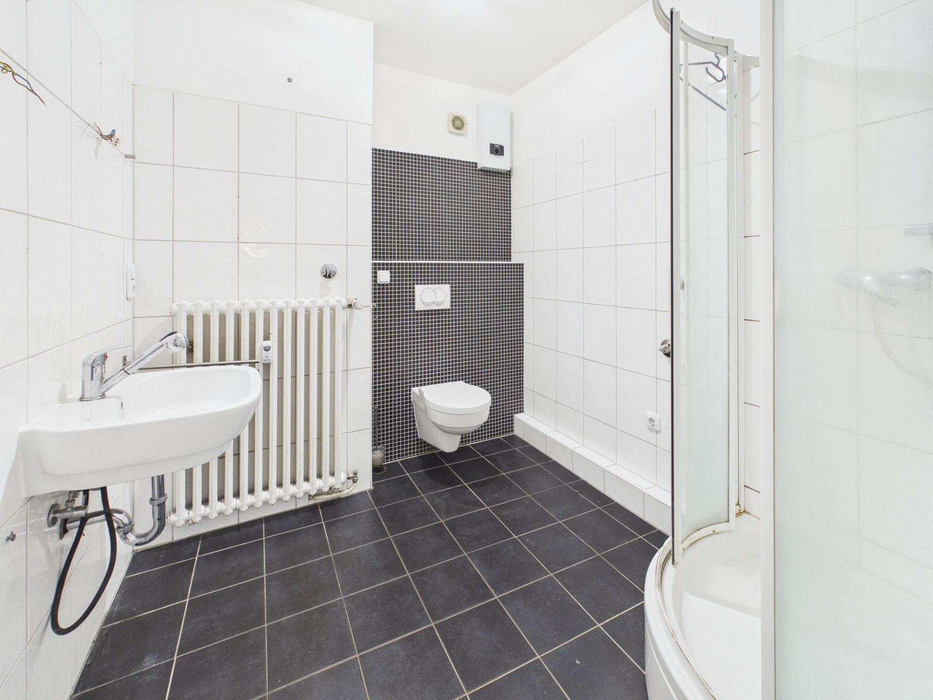 Modernes Badezimmer mit Dusche, Wandtoilette, Waschbecken und Heizkörper auf schwarzen Fliesen.