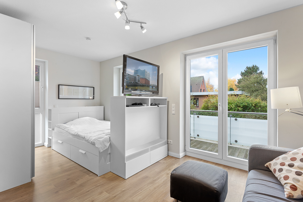Helles Schlafzimmer mit Bett, Sofa, Fernseher und großem Fenster mit Blick auf einen Balkon.