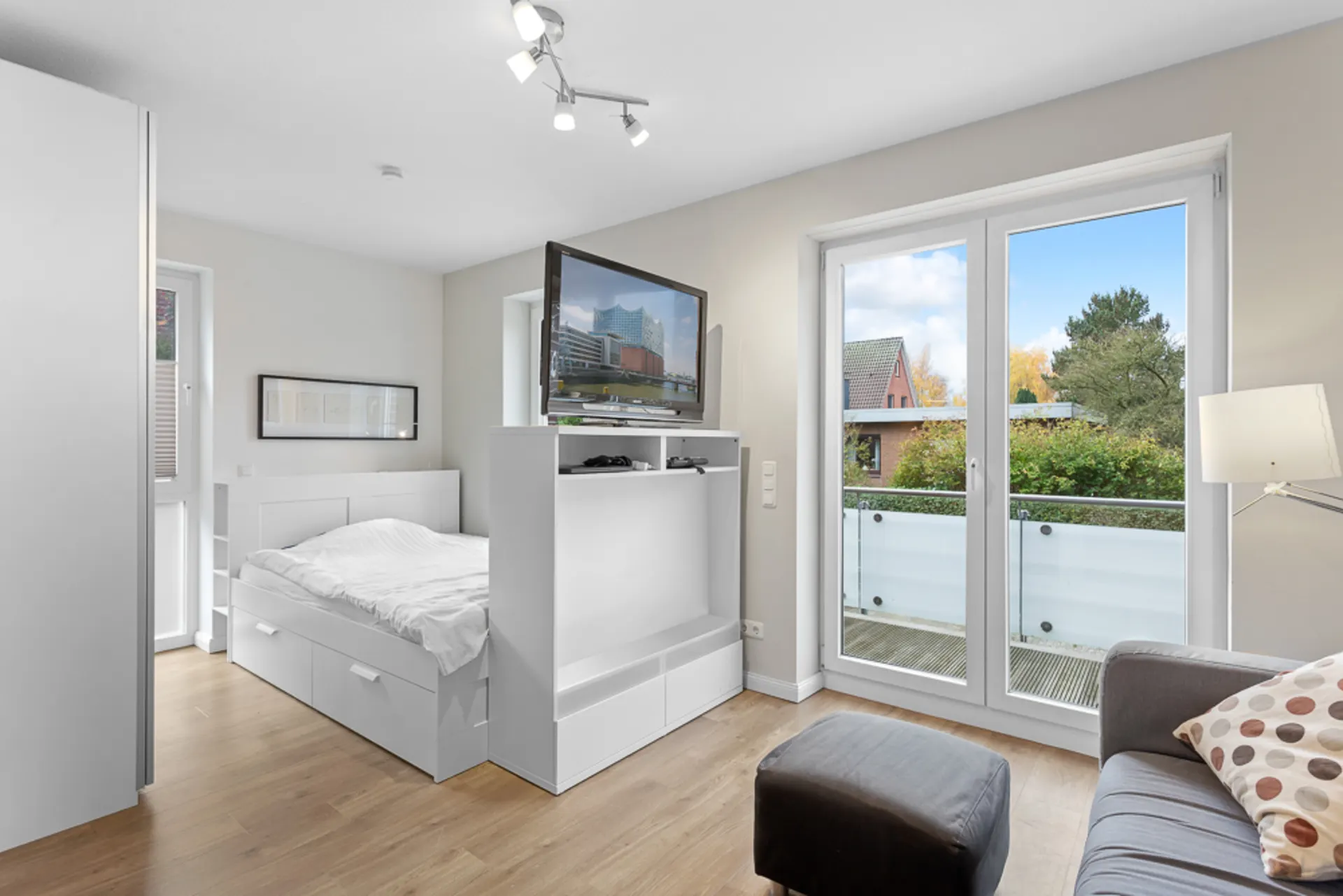 Helles Schlafzimmer mit Bett, Sofa, Fernseher und großem Fenster mit Blick auf einen Balkon.
