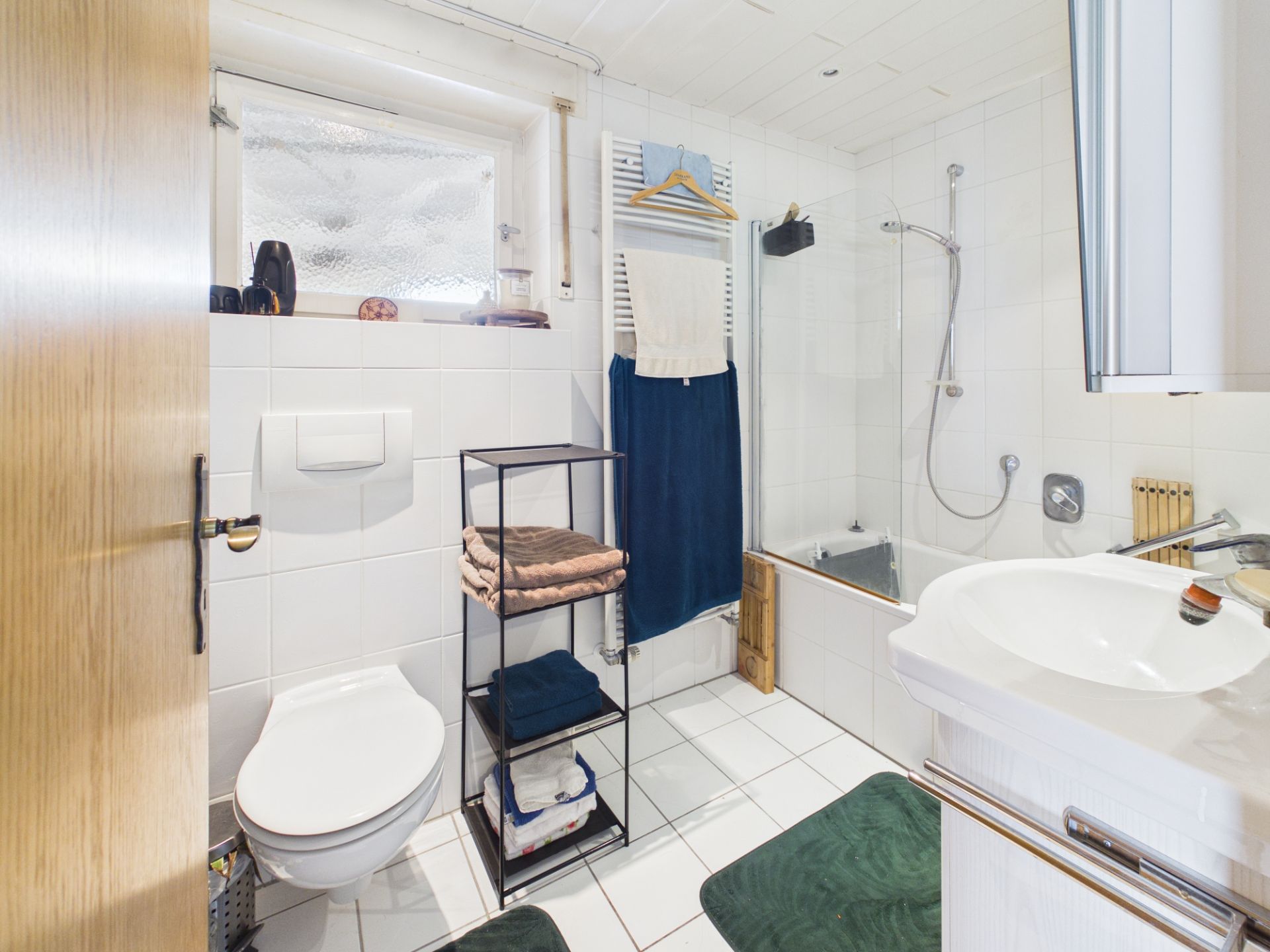 Helles Badezimmer mit WC, Waschbecken, Badewanne, Regal mit Handtüchern und Fenster.
