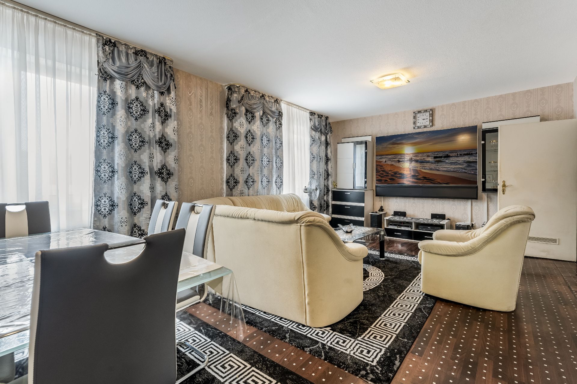 Wohnzimmer mit cremefarbenen Sofas, großem Fernseher, Esstisch und gemusterten Vorhängen.