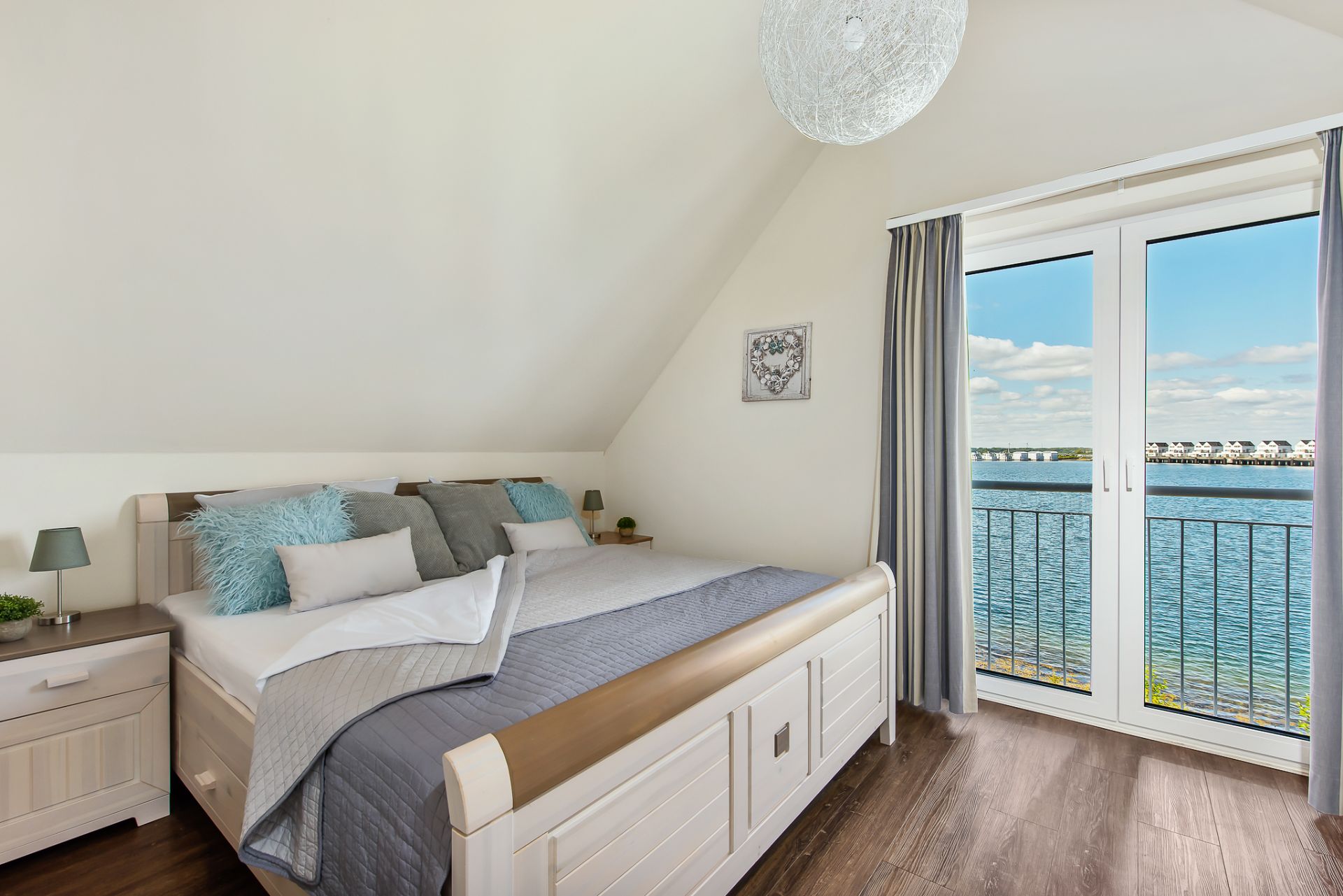 Helles Schlafzimmer mit Doppelbett, Nachtschränken und Balkonblick auf einen See.