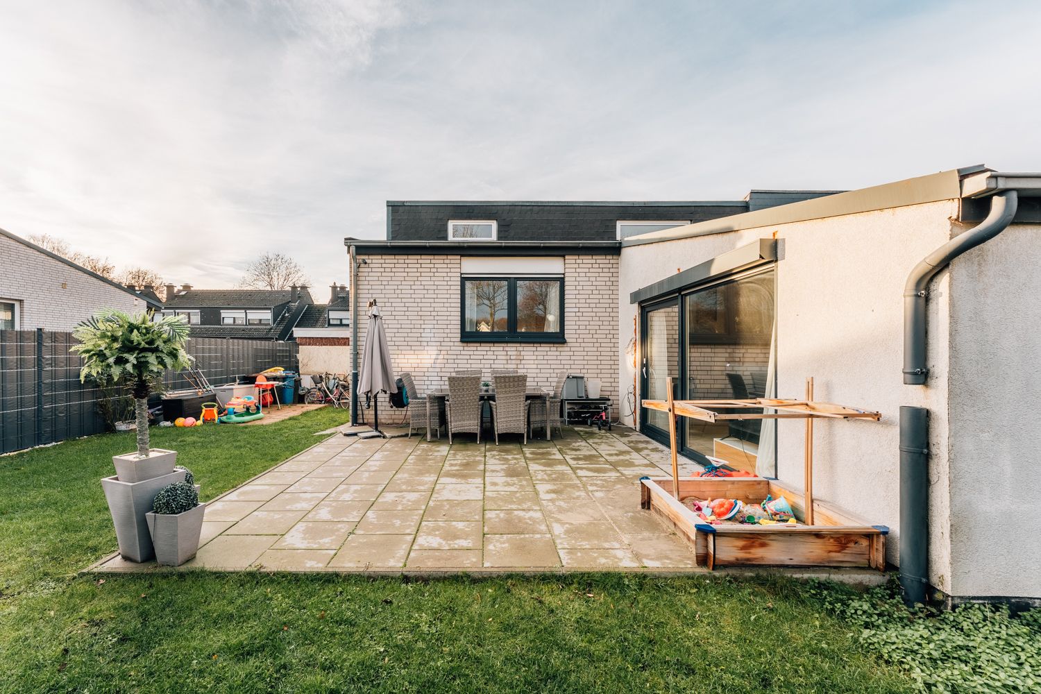 Garten mit Terrasse, Gartentisch, Stühlen, Spielzeug und Sandkasten vor einem Wohnhaus.