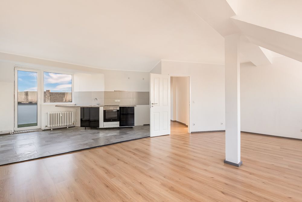 Helles, leeres Wohnzimmer mit offener Einbauküche, Parkettboden und großen Fenstern.