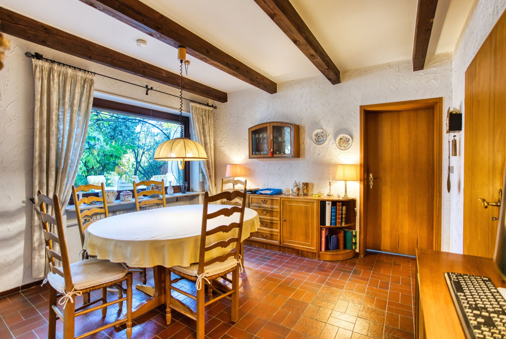 Esszimmer mit rundem Tisch, sechs Stühlen, Holzschrank, Fenster und sichtbaren Deckenbalken.