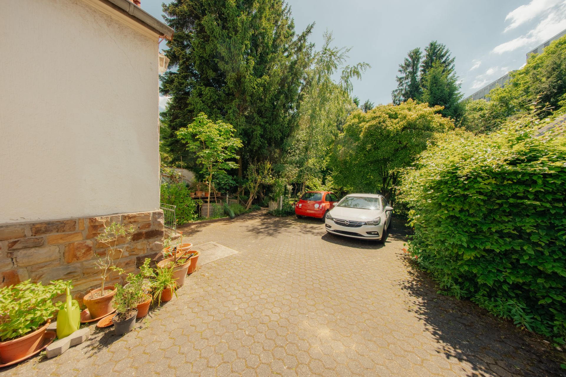 Gepflasterte Einfahrt mit zwei geparkten Autos, vielen Pflanzen und Bäumen neben einem Wohnhaus.