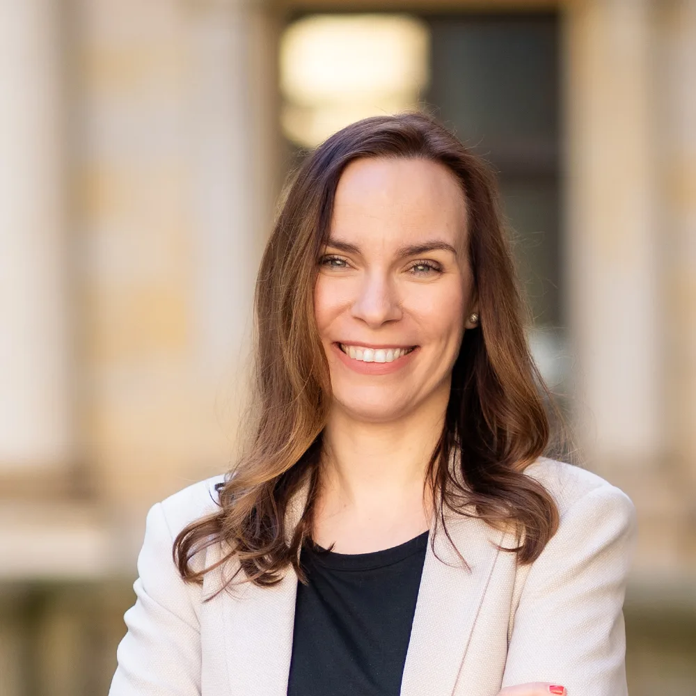 Frau mit langen braunen Haaren, hellem Blazer und verschränkten Armen lächelt draußen.