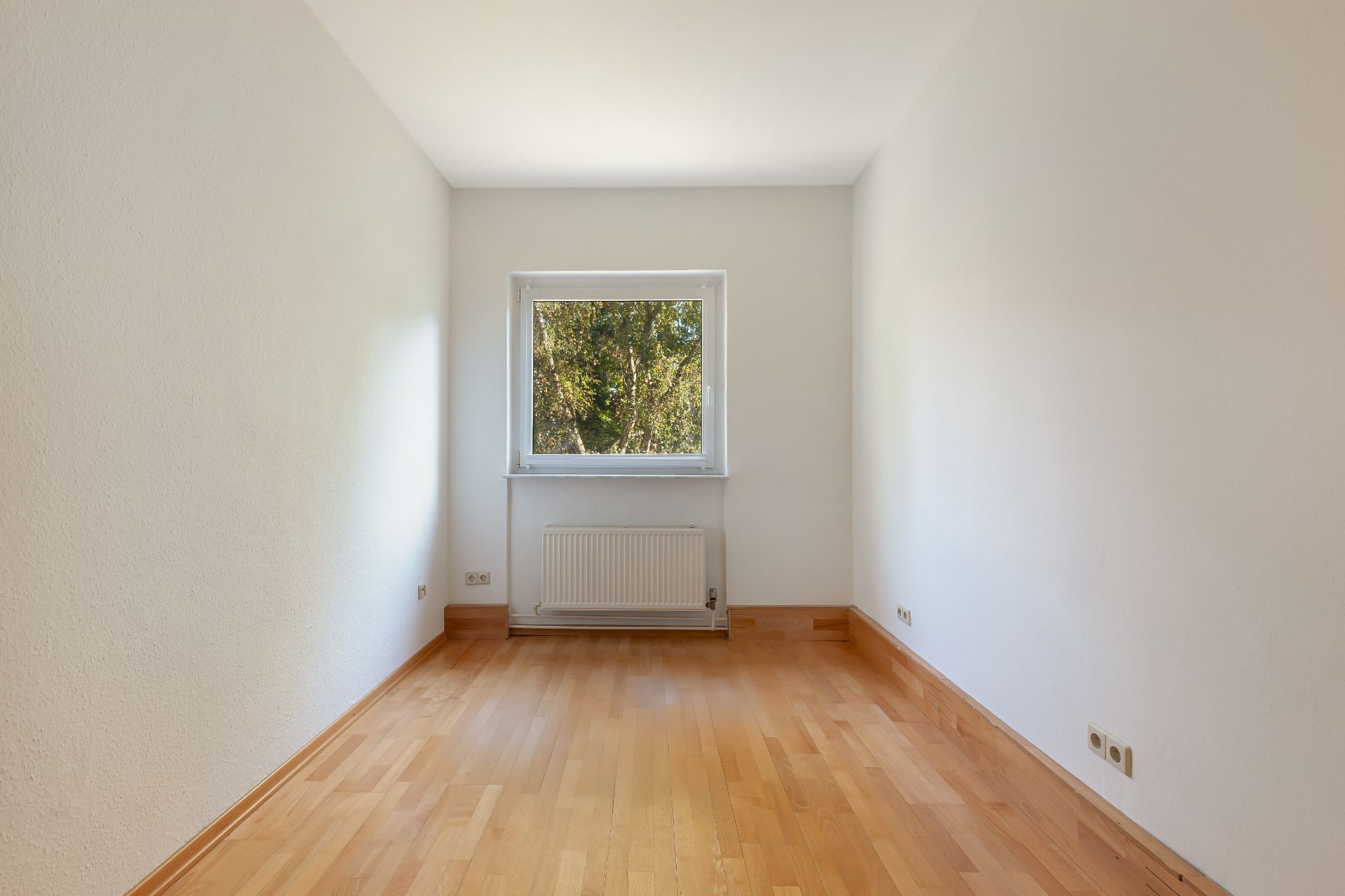 Leerer Raum mit Holzboden, weiß gestrichenen Wänden und einem Fenster mit Blick ins Grüne.