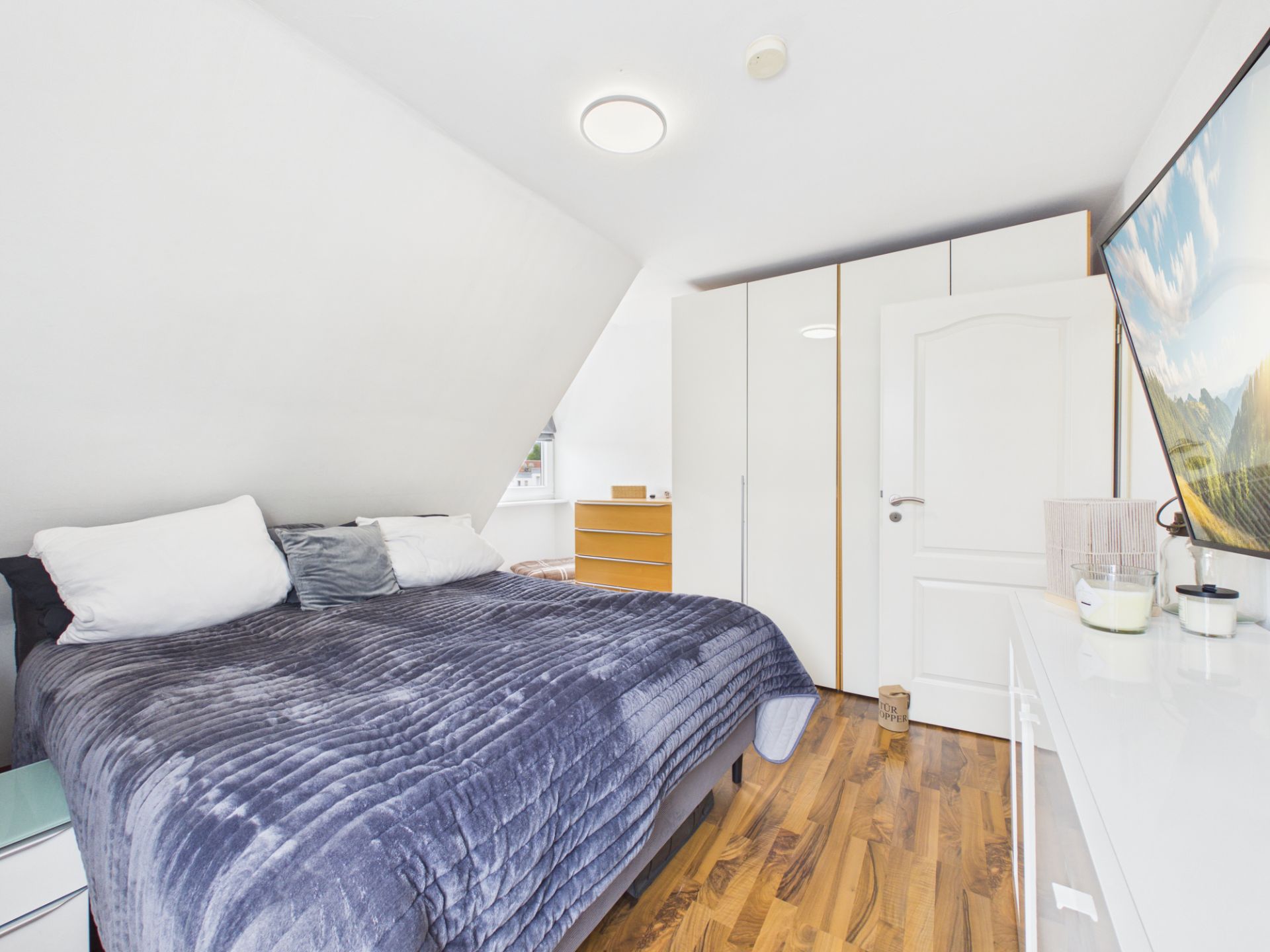 Helles Schlafzimmer mit Doppelbett, Kleiderschrank, Kommode und großem Fernseher an der Wand.