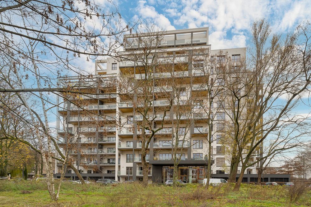 Mehrstöckiges Wohnhaus mit Balkonen, umgeben von Bäumen und grüner Wiese.