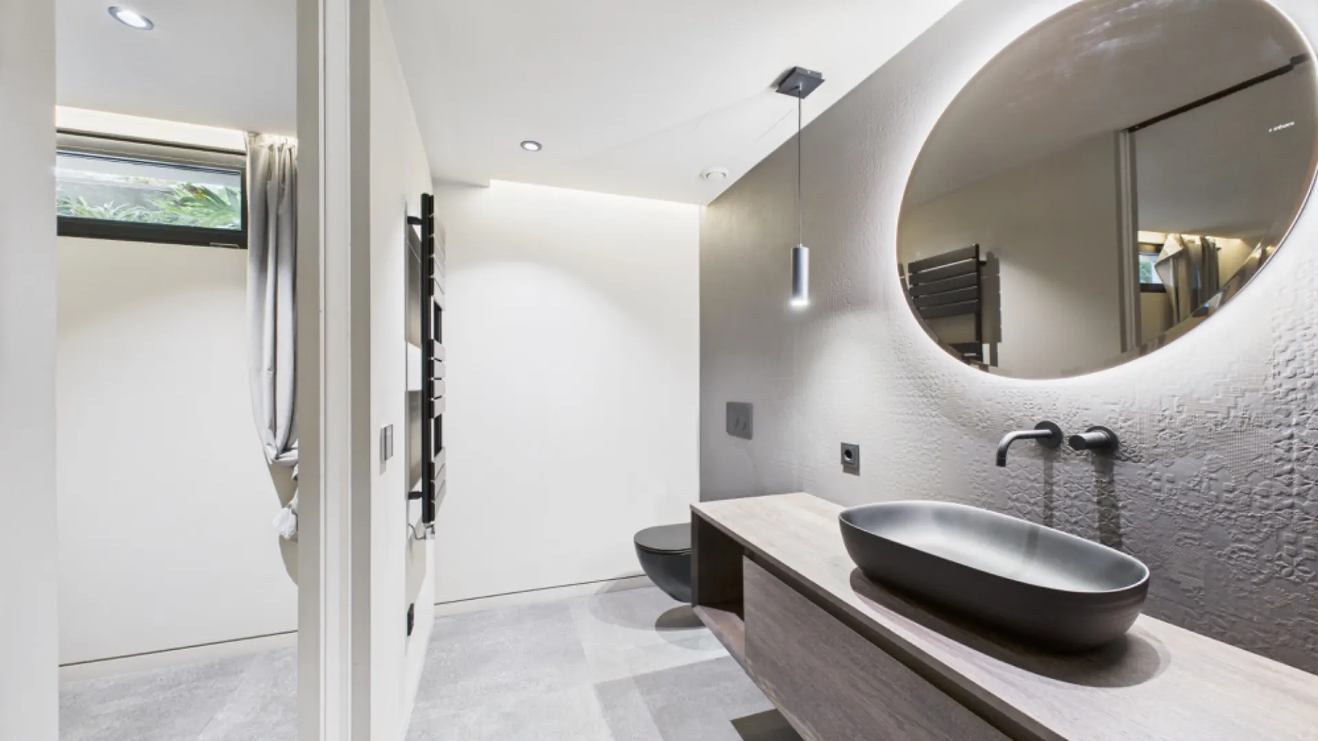 Modernes Badezimmer mit rundem Spiegel, schwarzem Waschbecken und heller Beleuchtung.