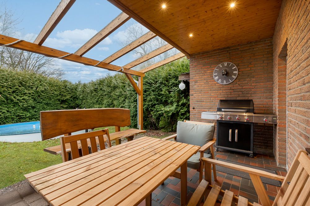 Überdachte Terrasse mit Holztisch, Stühlen, Grill, Uhr an der Wand und Blick auf den Garten.