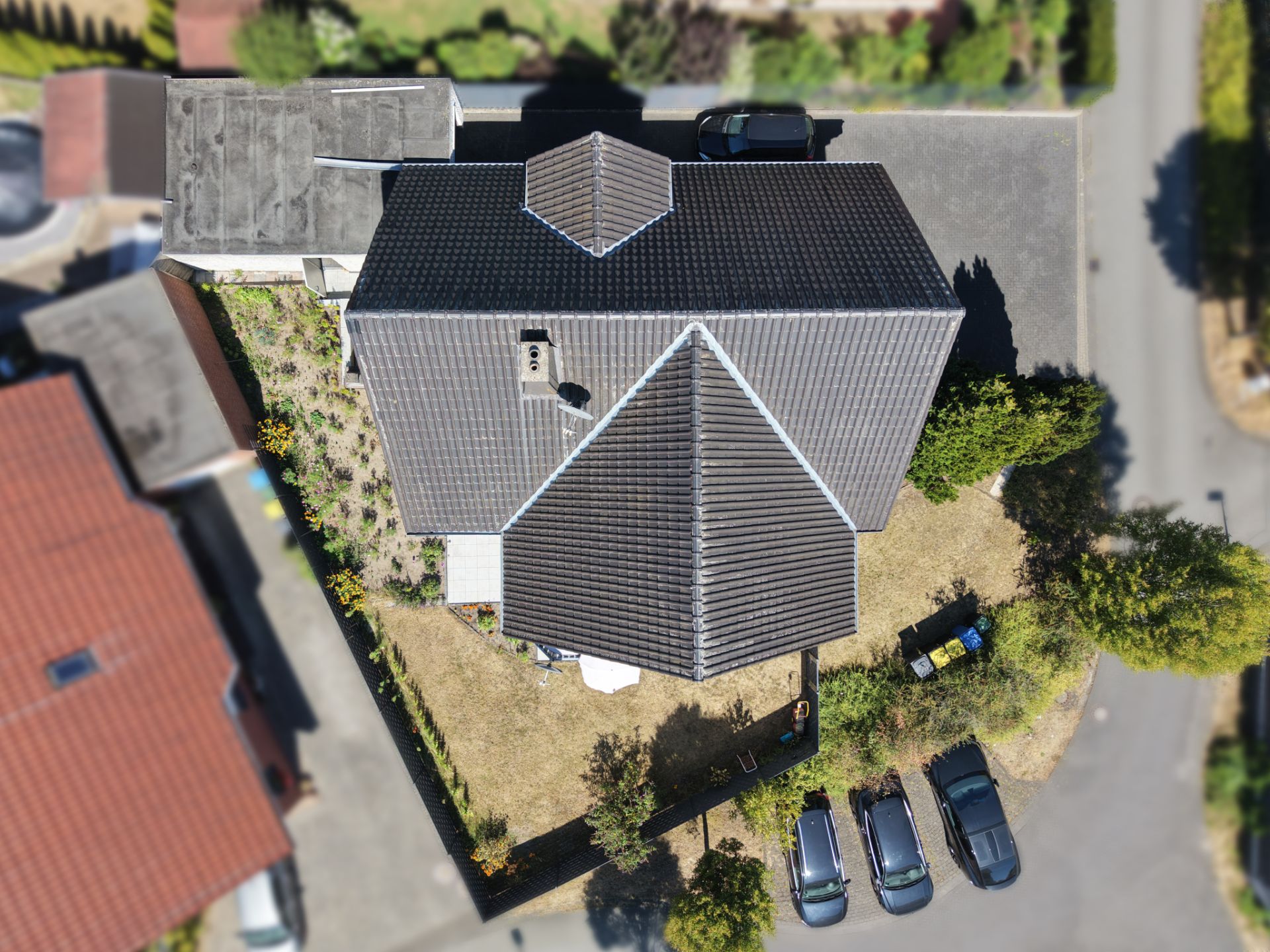 Luftbild eines Wohnhauses mit dunklem Dach, Garten und angrenzendem Parkplatz mit Autos.