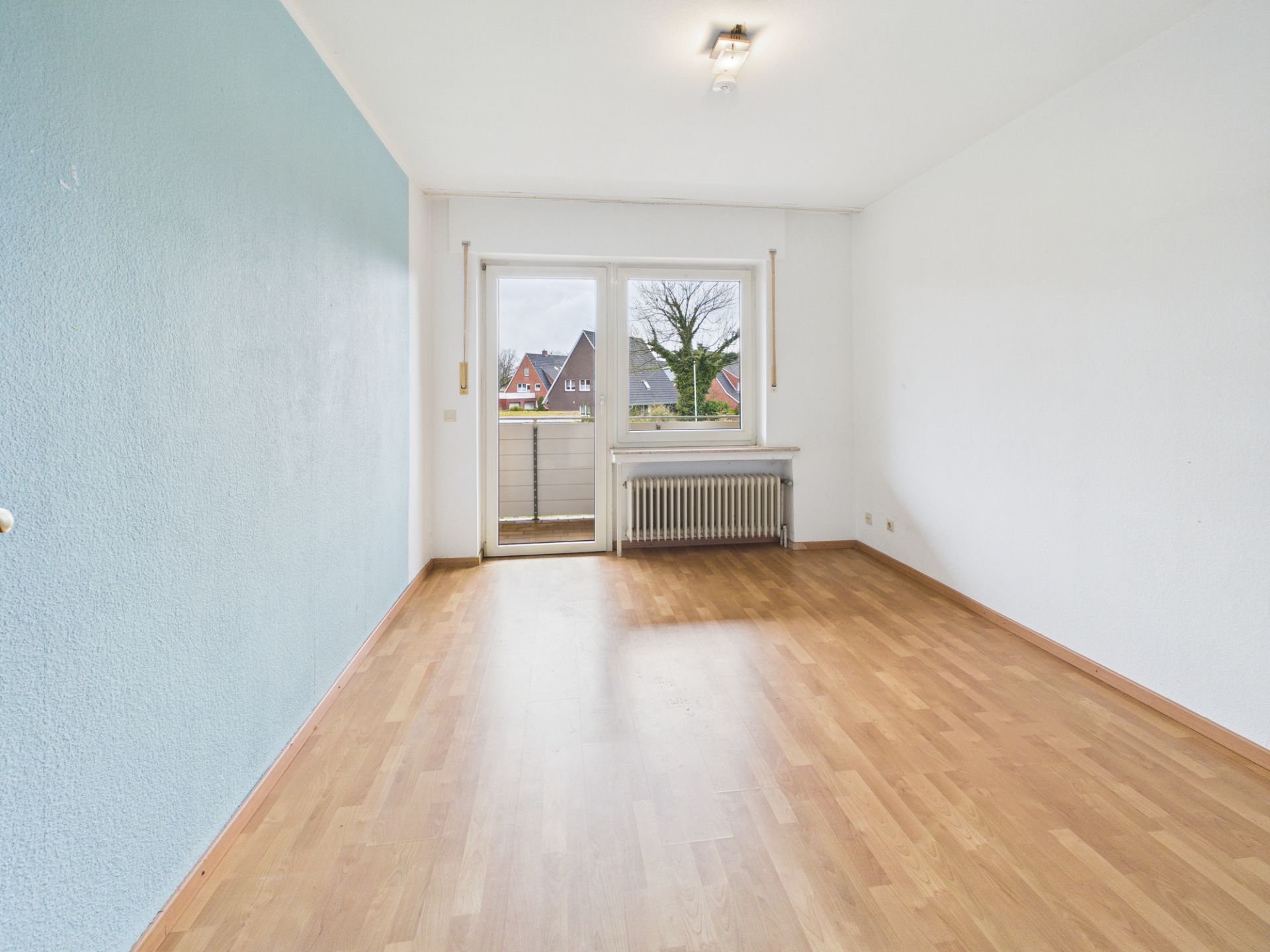 Leeres Zimmer mit Holzfußboden, blauer Wand, Heizung und Tür zu einem Balkon.
