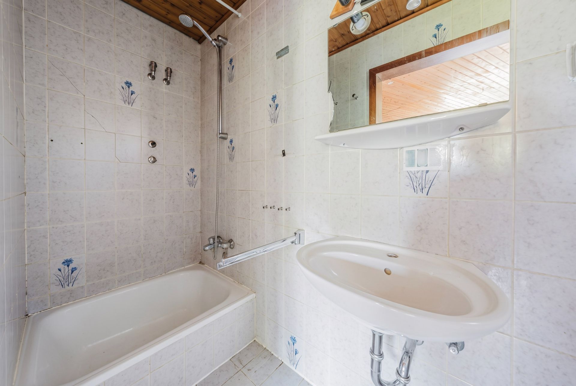 Kleines Badezimmer mit Badewanne, Waschbecken, Spiegel und hellen Fliesen mit Blumenmuster.