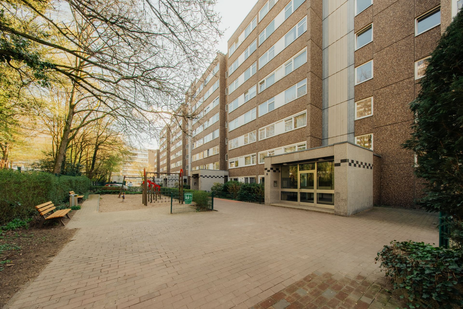 Mehrstöckiges Wohngebäude mit Spielplatz, Sitzbänken und Bäumen im Hof.