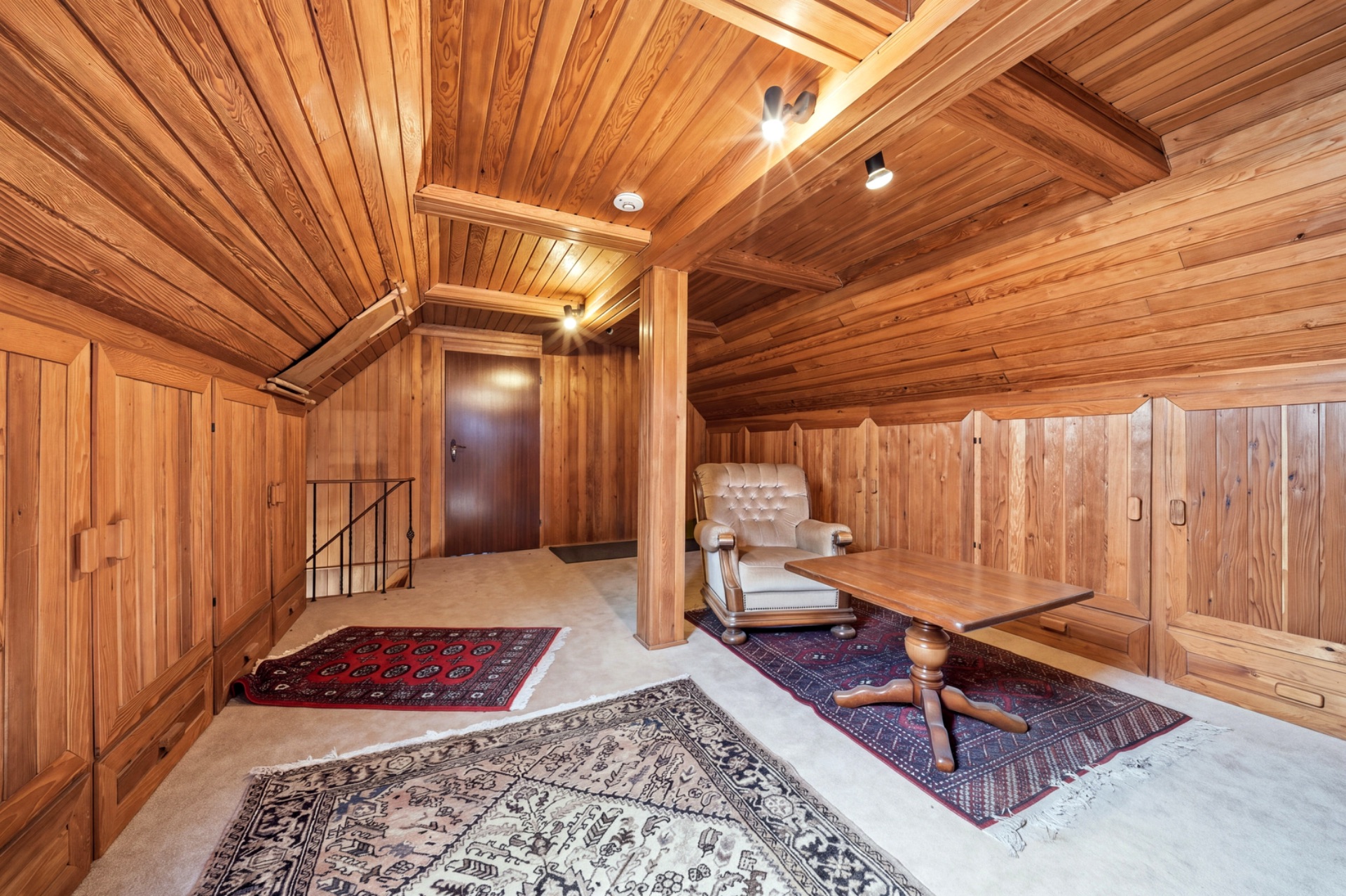 Dachzimmer mit Holzverkleidung, Sessel, Holztisch und Teppichen auf hellem Boden.