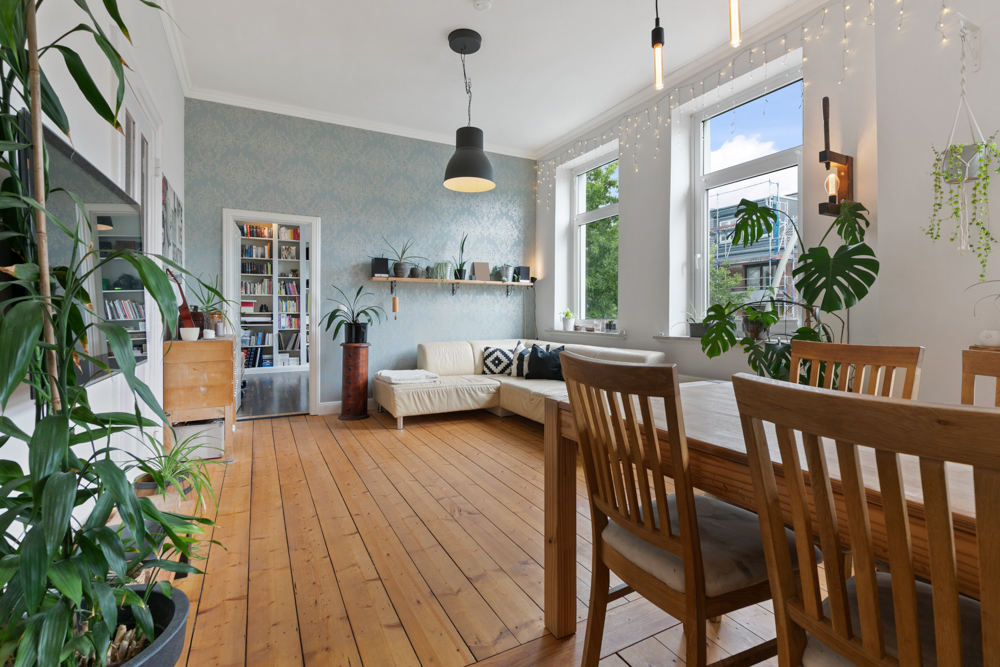 Helles Wohnzimmer mit Holzfußboden, Esstisch, Sofa, Pflanzen und großen Fenstern.