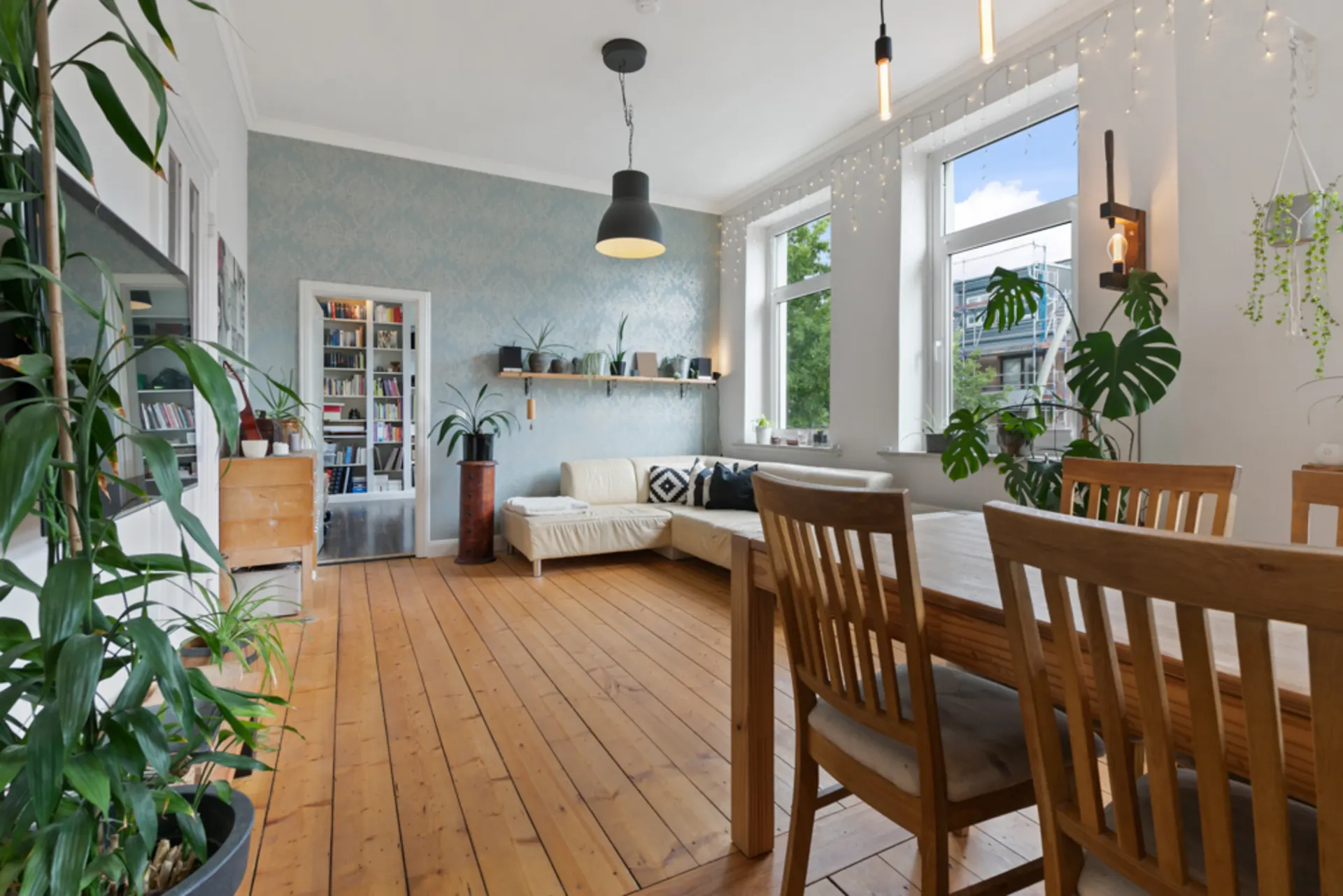 Helles Wohnzimmer mit Holzfußboden, Esstisch, Sofa, Pflanzen und großen Fenstern.