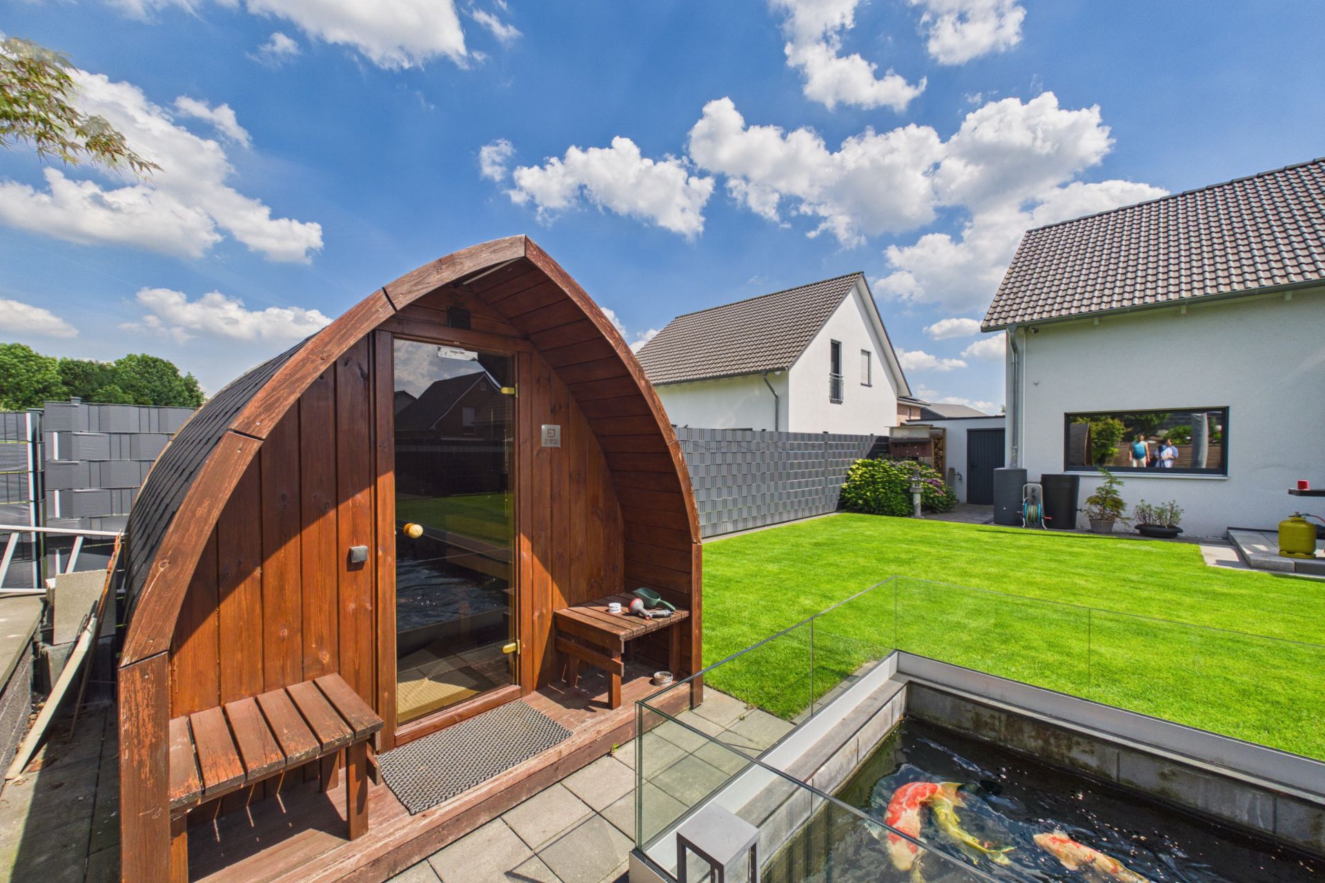 Garten mit Holzhütte, Sitzbank, Teich mit Fischen und Rasenfläche, im Hintergrund Wohnhäuser.