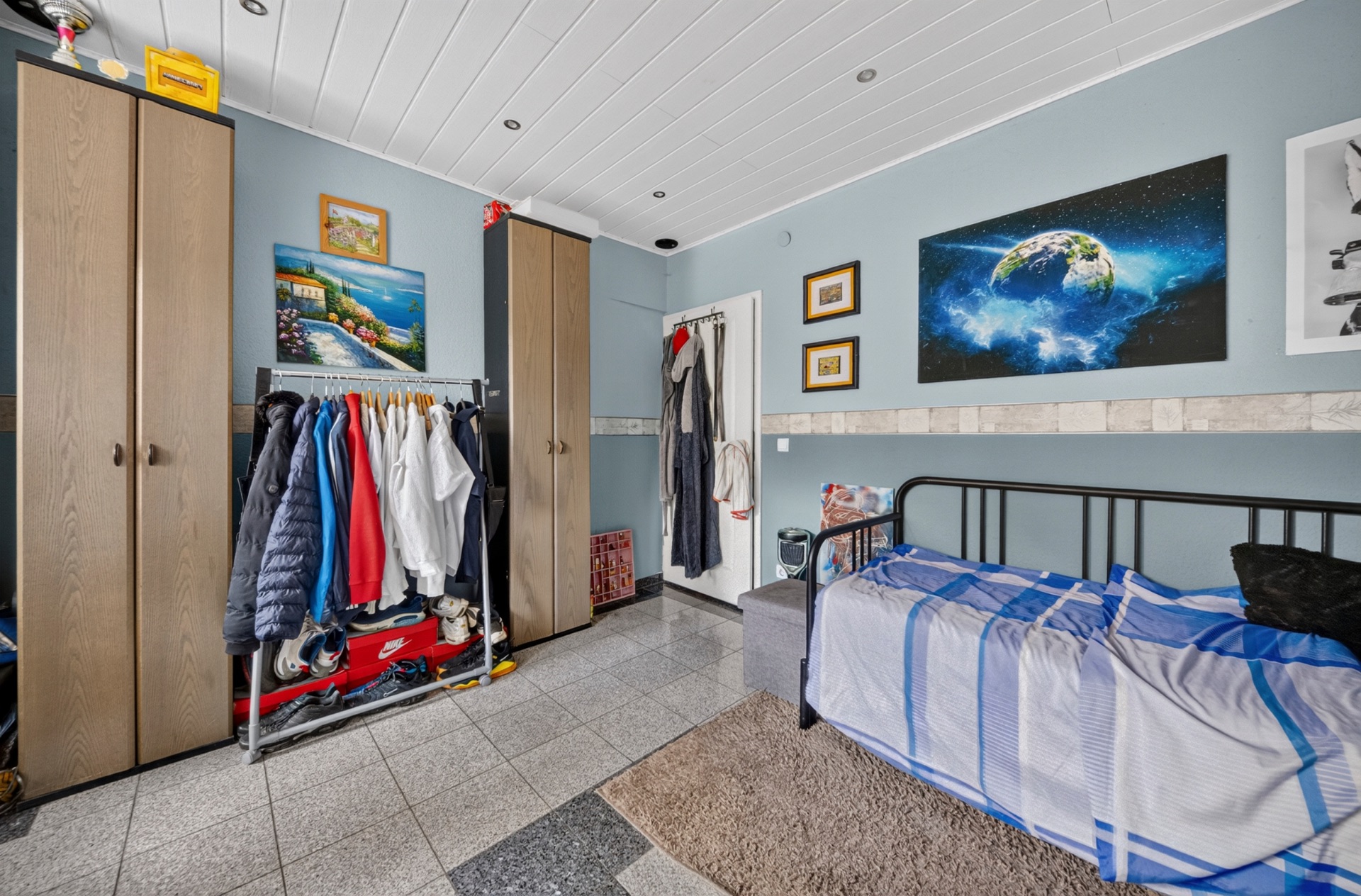 Zimmer mit zwei Schränken, einem Bett, Kleiderstange und Bildern an der blauen Wand.