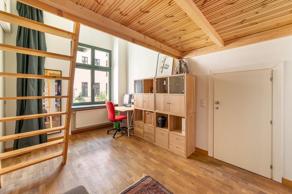Helles Zimmer mit Hochbett, Holzregal, Schreibtisch und rotem Stuhl neben großem Fenster.