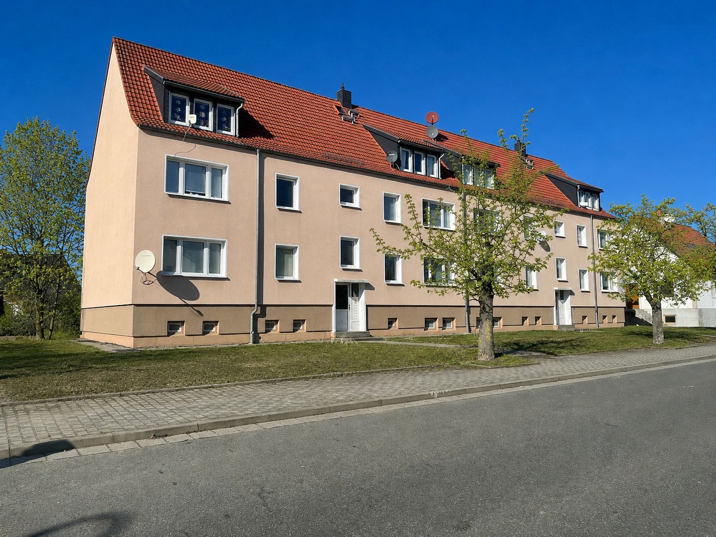 Dreistöckiges Wohnhaus mit rotem Dach, Bäumen davor und Gehweg an der Straße.